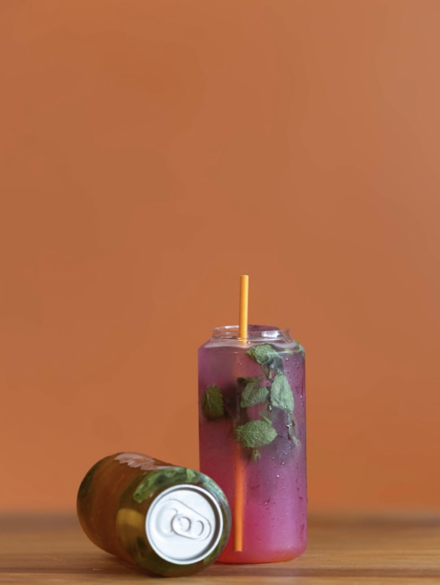 Pink iced drink with mint next to a can, on an orange backdrop