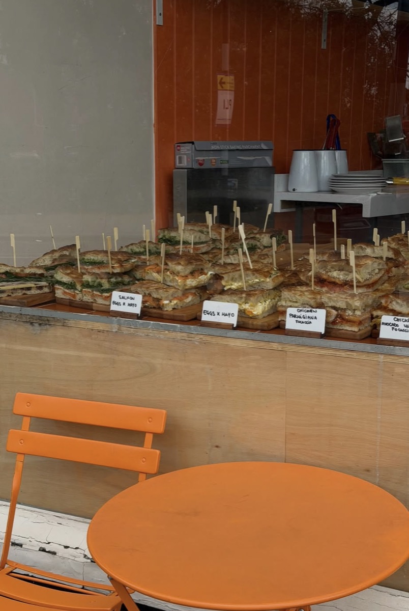 Focaccia sandwiches lined up on the counter, with an orange table and chair outside